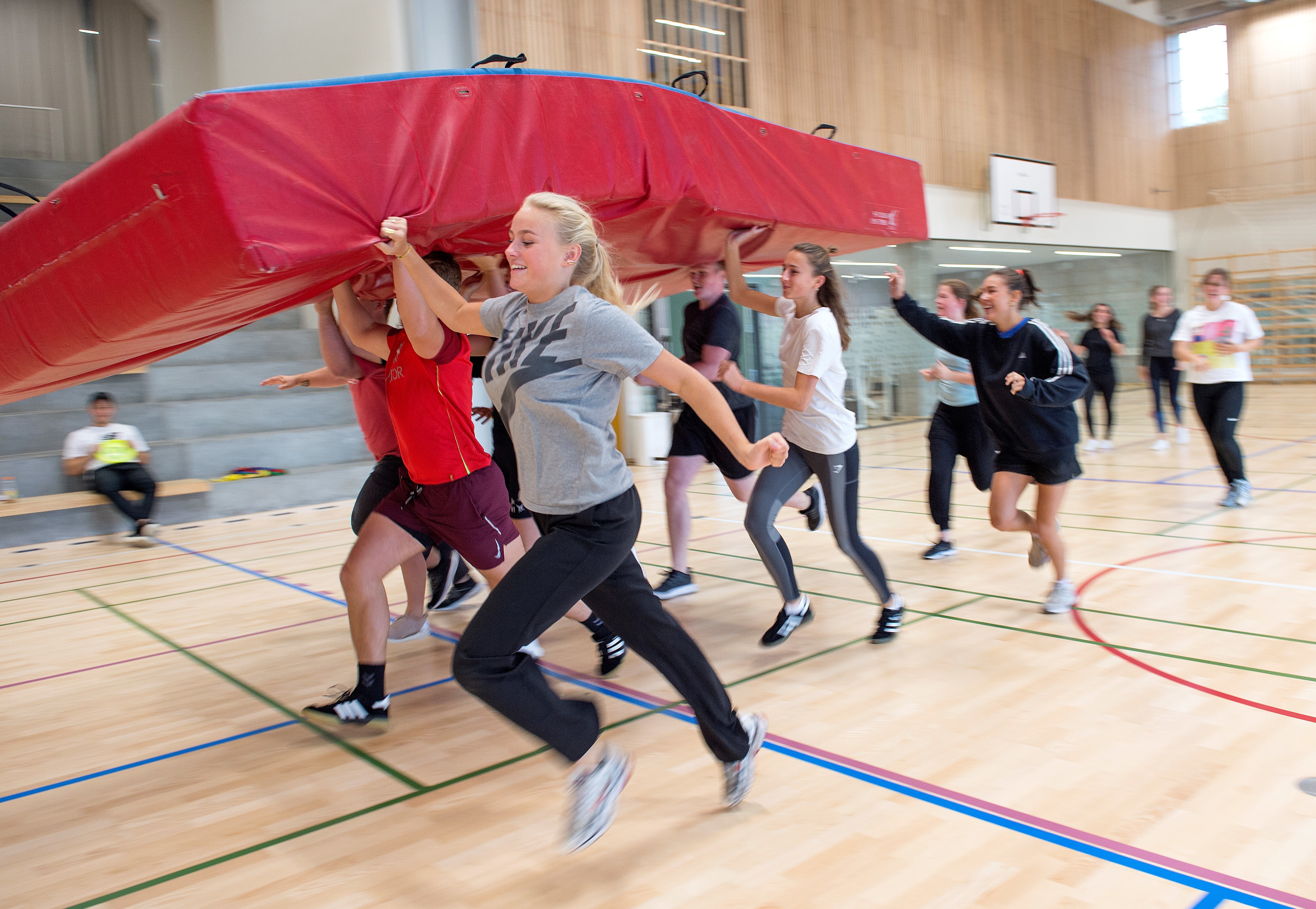 Cirka 10 elever har idræt i hallen. De løber i gruppe, mens de sammen bærer stor madras over hovedet